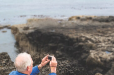 A senior couple sit on a blanket at the top of a hill looking over their family who are playing on the rocks by the sea below them. They are taking pictures of the horizon on a small camera.