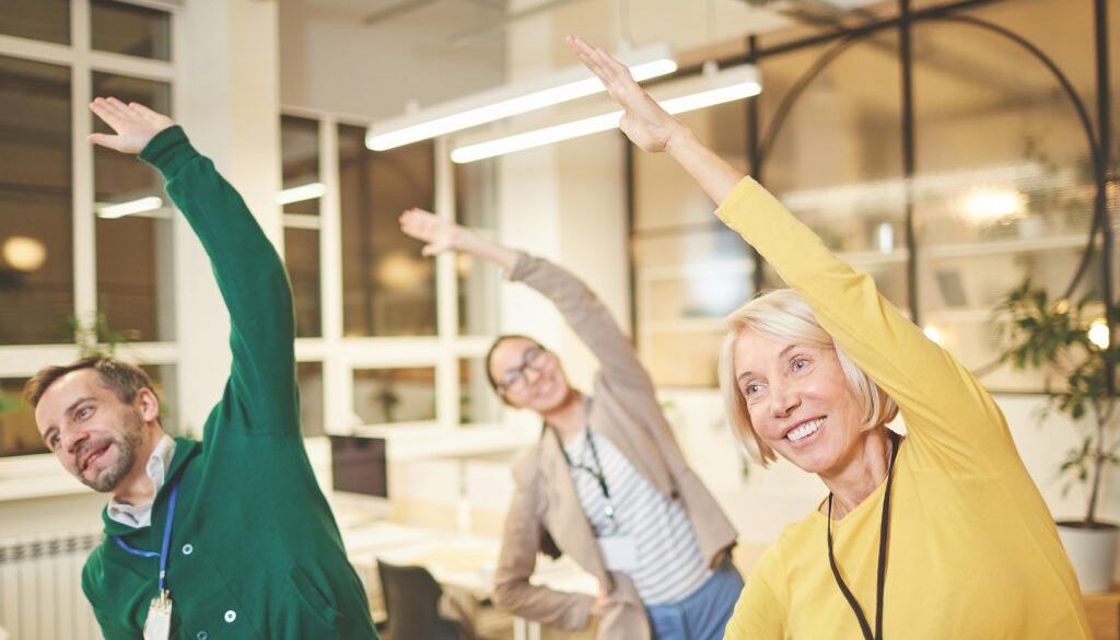 Cheerful active company employees in bright clothing exercising in office and making bending aside