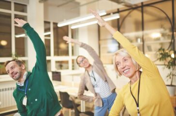 Cheerful active company employees in bright clothing exercising in office and making bending aside