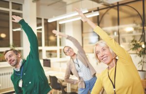 Healthy People Equals Healthy Profits Cheerful active company employees in bright clothing exercising in office and making bending aside