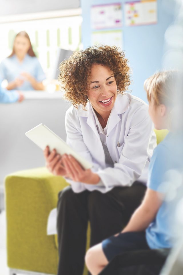 A female pharmacist sits with a male customer in the pharmacist consultation area and discusses his prescription and choice of medication viewing the details on a digital tablet. In the background a senior woman and granddaughter stand at the dispensing counter and are served by a female pharmacy assistant .