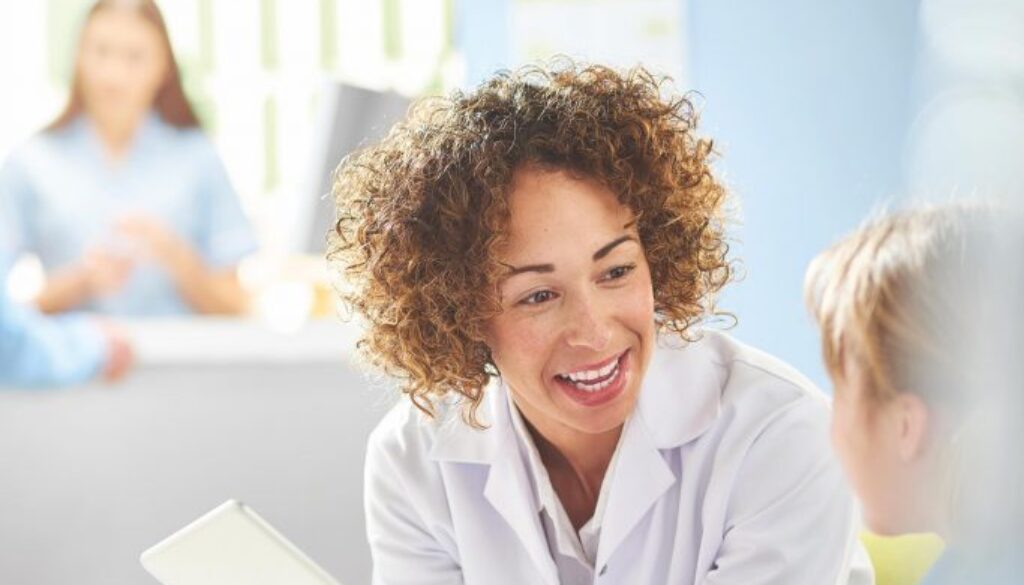 A female pharmacist sits with a male customer in the pharmacist consultation area and discusses his prescription and choice of medication viewing the details on a digital tablet. In the background a senior woman and granddaughter stand at the dispensing counter and are served by a female pharmacy assistant .