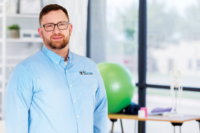 Cheerful mid adult physical therapist smiles confidently in a gym. She is holding hand weights.