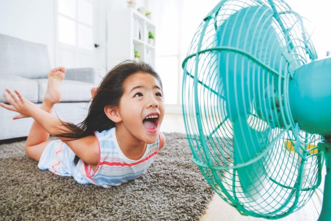 Don’t Sweat The Heat sweet beautiful little children lying down on living room floor and face to electric fan enjoying cool wind with flying posing with selective focus photo.