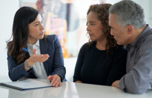 Are You Fully Prepared For Your Golden Years? An adviser meets with a mixed race middle-aged couple to discuss their life insurance or financial needs. The couple is dressed casually and the adviser is in a suit. They are seated around the clients kitchen table.