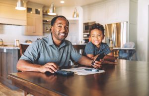Break The Cycle Of Financial Illiteracy A middle-aged man sits at his dining room table with his young son teaching him about home finances. He is working from home and telecommuting.