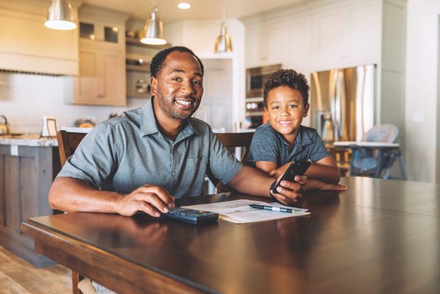 A middle-aged man sits at his dining room table with his young son teaching him about home finances. He is working from home and telecommuting.