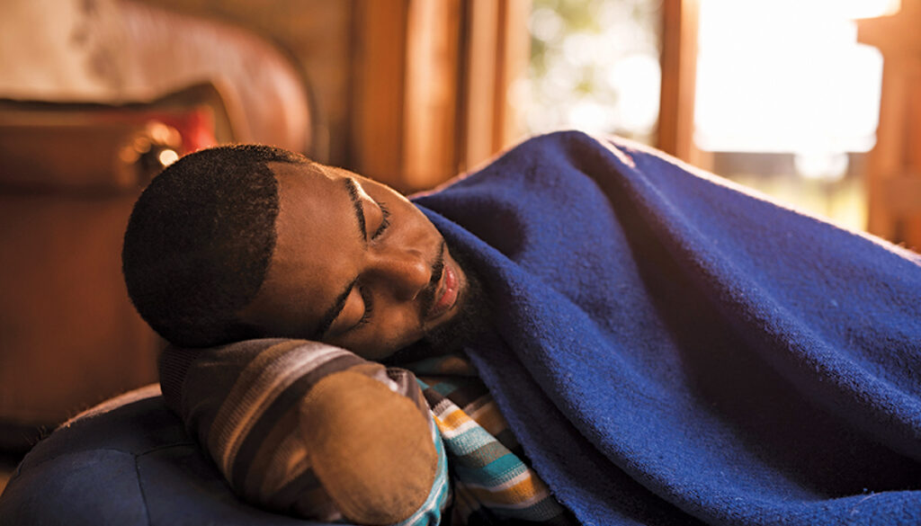 Tired black man lying on a bean bag and sleeping while being covered with blue blanket.
