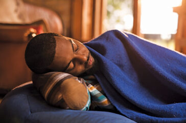 Tired black man lying on a bean bag and sleeping while being covered with blue blanket.