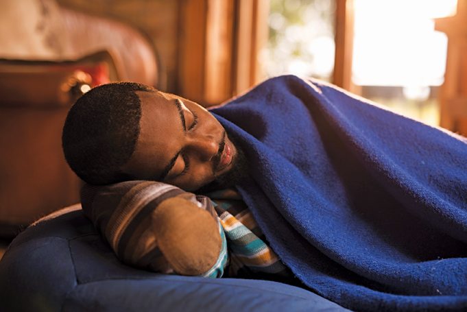 Take A Nap Tired black man lying on a bean bag and sleeping while being covered with blue blanket.