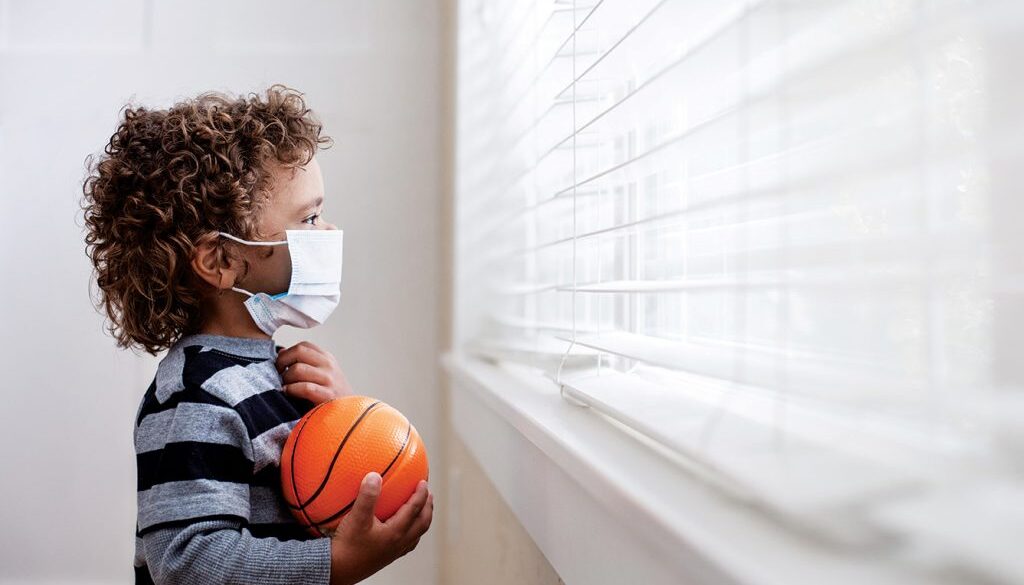 A young boy looking out the window wearing a protective facemark while seeking protection from COVID-19, or the novel coronavirus, by sheltering in place in his home.