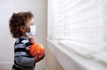 A young boy looking out the window wearing a protective facemark while seeking protection from COVID-19, or the novel coronavirus, by sheltering in place in his home.