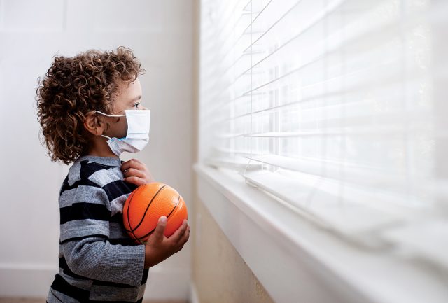 A young boy looking out the window wearing a protective facemark while seeking protection from COVID-19, or the novel coronavirus, by sheltering in place in his home.