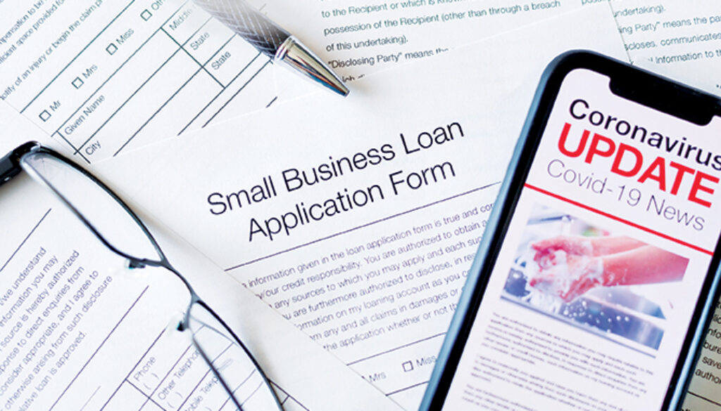 Small business loan application form. Close up of a mobile phone with Coronavirus covid-19 news update. There is a small business loan application in the background on the desk. The phone has an image of hand washing hygiene.