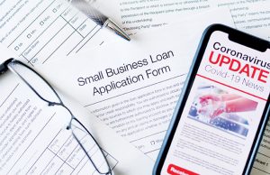 Tips For Small Business Owners During COVID-19 Small business loan application form. Close up of a mobile phone with Coronavirus covid-19 news update. There is a small business loan application in the background on the desk. The phone has an image of hand washing hygiene.
