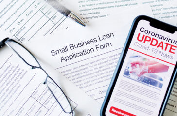 Small business loan application form. Close up of a mobile phone with Coronavirus covid-19 news update. There is a small business loan application in the background on the desk. The phone has an image of hand washing hygiene.