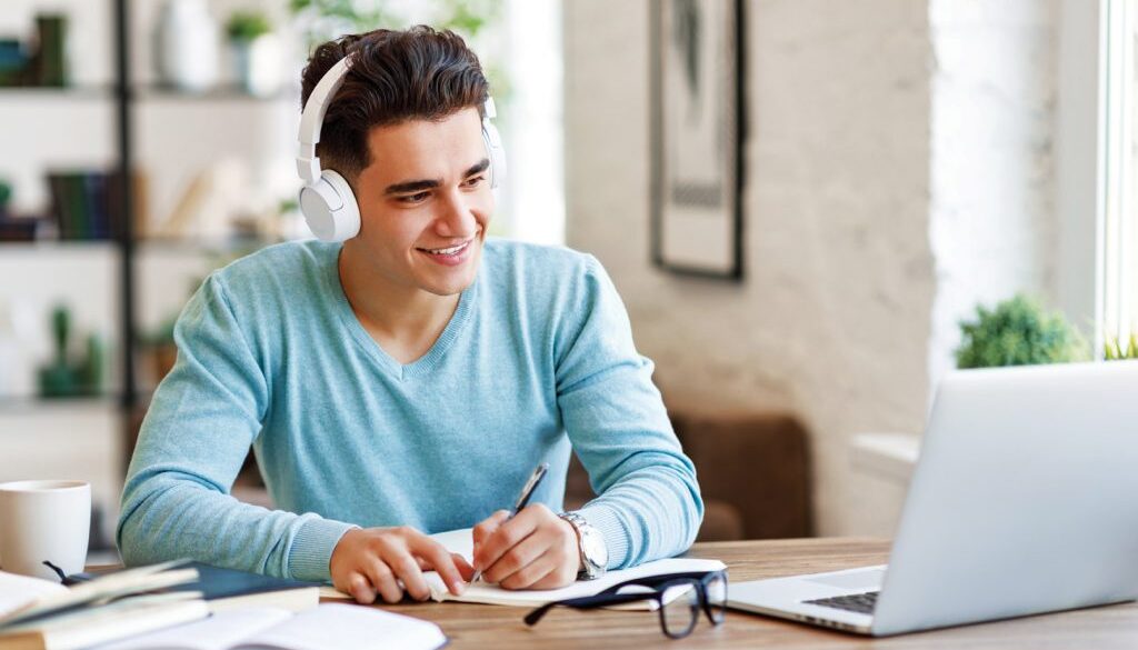Happy ethnic guy in headphones smiling and writing in notebook while sitting at table and listening to teacher during online lecture at home