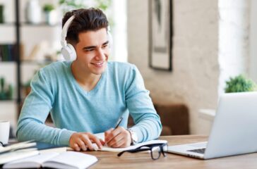 Happy ethnic guy in headphones smiling and writing in notebook while sitting at table and listening to teacher during online lecture at home
