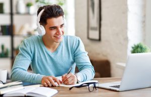 Moore Tutoring Happy ethnic guy in headphones smiling and writing in notebook while sitting at table and listening to teacher during online lecture at home