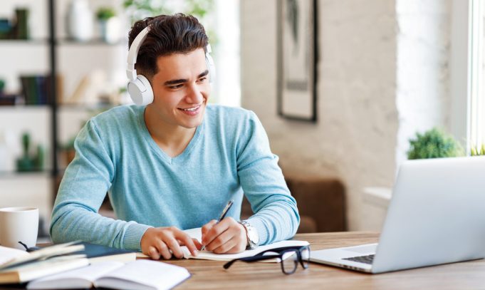 Moore Tutoring Happy ethnic guy in headphones smiling and writing in notebook while sitting at table and listening to teacher during online lecture at home