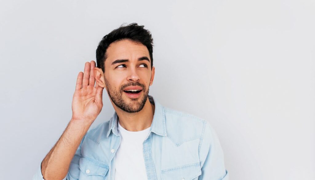 Portrait of handsome interested male placing hand on ear asking someone to speak up, isolated over white background. Young stylish man which overhears conversation in the studio. Copy space for text.