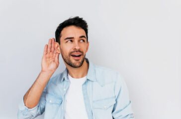 Portrait of handsome interested male placing hand on ear asking someone to speak up, isolated over white background. Young stylish man which overhears conversation in the studio. Copy space for text.