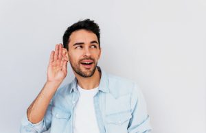Listen Up! Portrait of handsome interested male placing hand on ear asking someone to speak up, isolated over white background. Young stylish man which overhears conversation in the studio. Copy space for text.