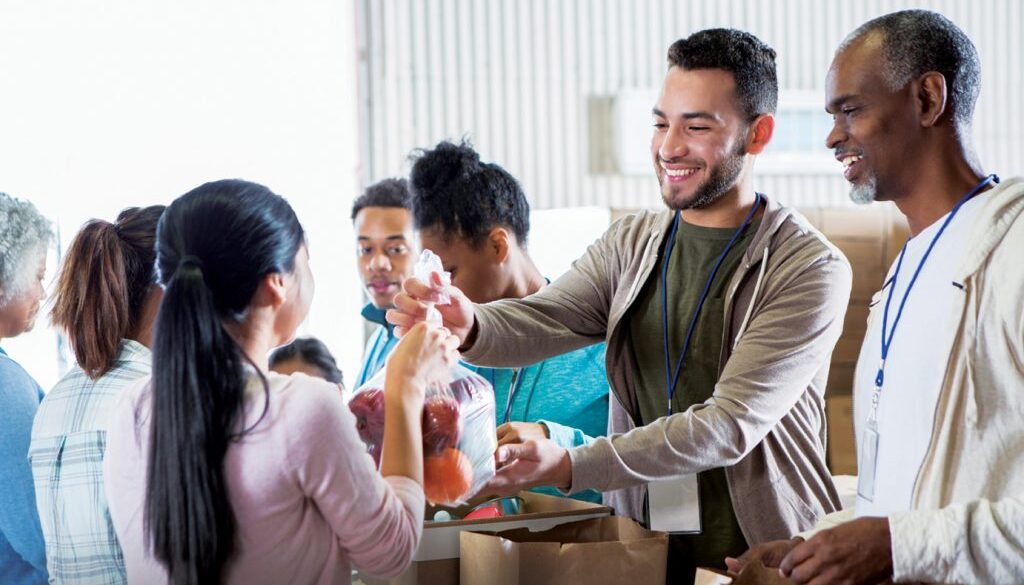 Young woman hands a bag of fruit to a young male food bank volunteer. The volunteers are receiving donations from their community to benefit a food bank.