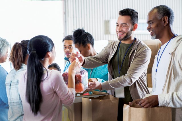 Young woman hands a bag of fruit to a young male food bank volunteer. The volunteers are receiving donations from their community to benefit a food bank.