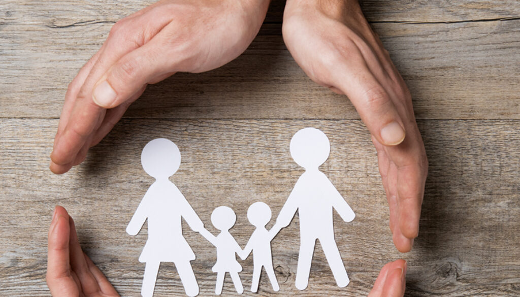 Close up of female and male hands protecting a paper chain family. Top view of two hands form a circle around white paper chain family on wooden table. Family care, insurance and helping hand concept.