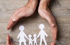 When Disaster Strikes… Close up of female and male hands protecting a paper chain family. Top view of two hands form a circle around white paper chain family on wooden table. Family care, insurance and helping hand concept.