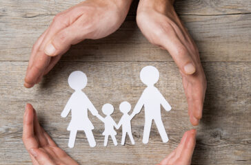 Close up of female and male hands protecting a paper chain family. Top view of two hands form a circle around white paper chain family on wooden table. Family care, insurance and helping hand concept.