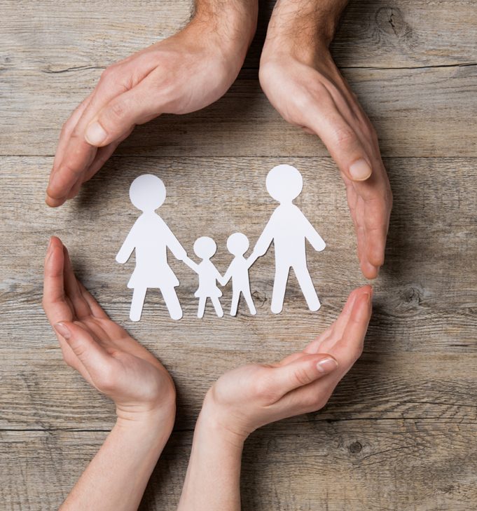 When Disaster Strikes… Close up of female and male hands protecting a paper chain family. Top view of two hands form a circle around white paper chain family on wooden table. Family care, insurance and helping hand concept.