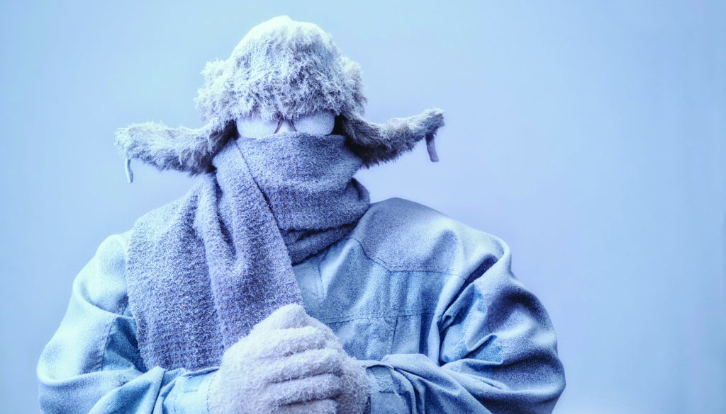 A frozen man with his hands clasped all bundled up in a fur trappers hat, scarf, parka and knit gloves, covered in snow and frost trying to stay warm on a very cold gray Winter's day.