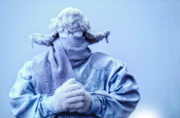A frozen man with his hands clasped all bundled up in a fur trappers hat, scarf, parka and knit gloves, covered in snow and frost trying to stay warm on a very cold gray Winter's day.