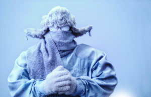 Thawing Out A frozen man with his hands clasped all bundled up in a fur trappers hat, scarf, parka and knit gloves, covered in snow and frost trying to stay warm on a very cold gray Winter's day.