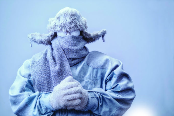 Thawing Out A frozen man with his hands clasped all bundled up in a fur trappers hat, scarf, parka and knit gloves, covered in snow and frost trying to stay warm on a very cold gray Winter's day.