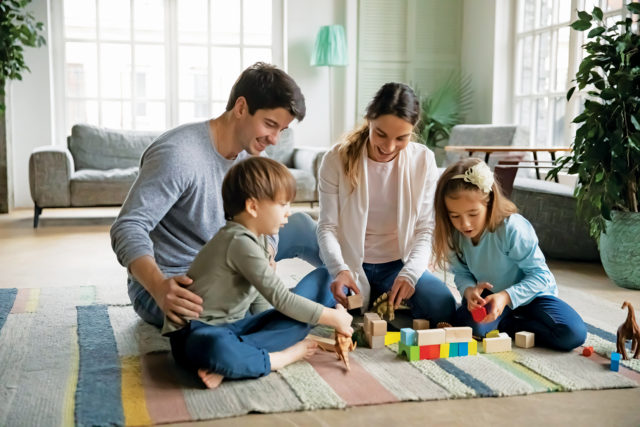 Affectionate caring young parents sitting on floor carpet with little joyful children siblings, playing toys together at home. Happy caucasian family with small kids involved in weekend game indoors.