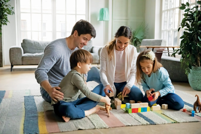 Siblings In Foster Care are Better Together Affectionate caring young parents sitting on floor carpet with little joyful children siblings, playing toys together at home. Happy caucasian family with small kids involved in weekend game indoors.