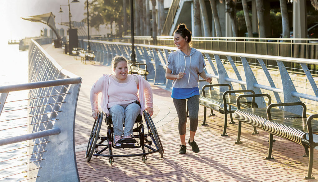 Two friends exercising along a city waterfront. The young woman in the wheelchair has spina bifida. Her friend jogging beside her and talking is a mid adult Hispanic woman in her 30s.