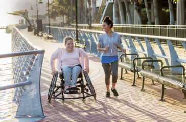 Two friends exercising along a city waterfront. The young woman in the wheelchair has spina bifida. Her friend jogging beside her and talking is a mid adult Hispanic woman in her 30s.