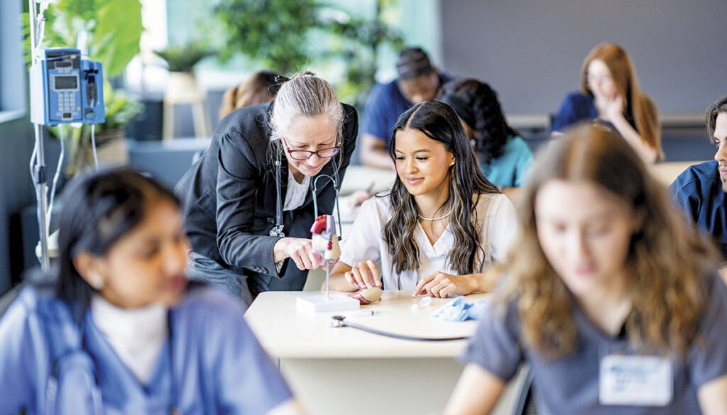 A large group of medical students are seen sitting in rows of desk as they work in pairs on various activities. They are each dressed professionally in scrubs and are working diligently on perfecting their skills. Their professor is seen checking in on various students in the background.