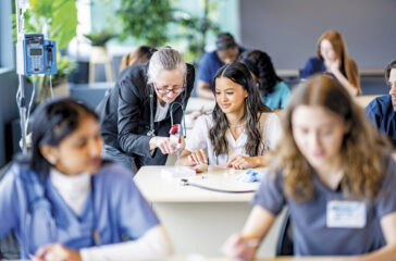 A large group of medical students are seen sitting in rows of desk as they work in pairs on various activities. They are each dressed professionally in scrubs and are working diligently on perfecting their skills. Their professor is seen checking in on various students in the background.