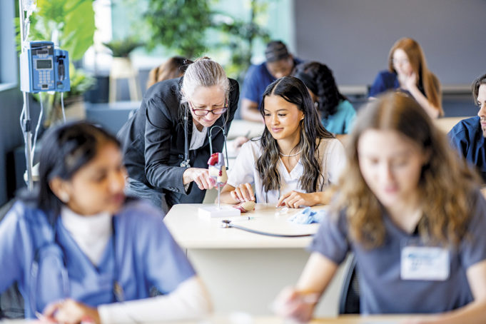 Wellstar Supports Developing Future Healthcare Professionals at Cobb Innovation Technology Academy A large group of medical students are seen sitting in rows of desk as they work in pairs on various activities. They are each dressed professionally in scrubs and are working diligently on perfecting their skills. Their professor is seen checking in on various students in the background.