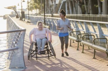 Two friends exercising along a city waterfront. The young woman in the wheelchair has spina bifida. Her friend jogging beside her and talking is a mid adult Hispanic woman in her 30s.