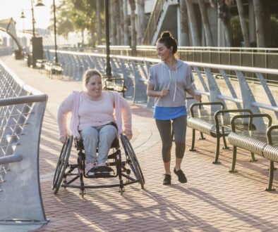 Two friends exercising along a city waterfront. The young woman in the wheelchair has spina bifida. Her friend jogging beside her and talking is a mid adult Hispanic woman in her 30s.