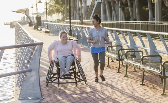 Two friends exercising along a city waterfront. The young woman in the wheelchair has spina bifida. Her friend jogging beside her and talking is a mid adult Hispanic woman in her 30s.