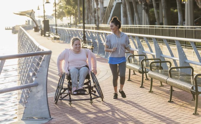 How Walkability and Movability Can Impact People, Place, and Prosperity in Cobb County Two friends exercising along a city waterfront. The young woman in the wheelchair has spina bifida. Her friend jogging beside her and talking is a mid adult Hispanic woman in her 30s.