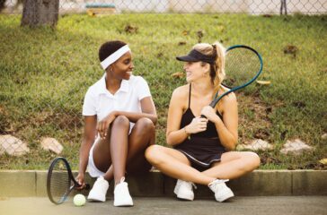 Young beautiful women wearing tennis clothes, sitting on the floor of a tennis court and chatting just before to start a tennis game. Sportswomen getting ready for a tennis match.
