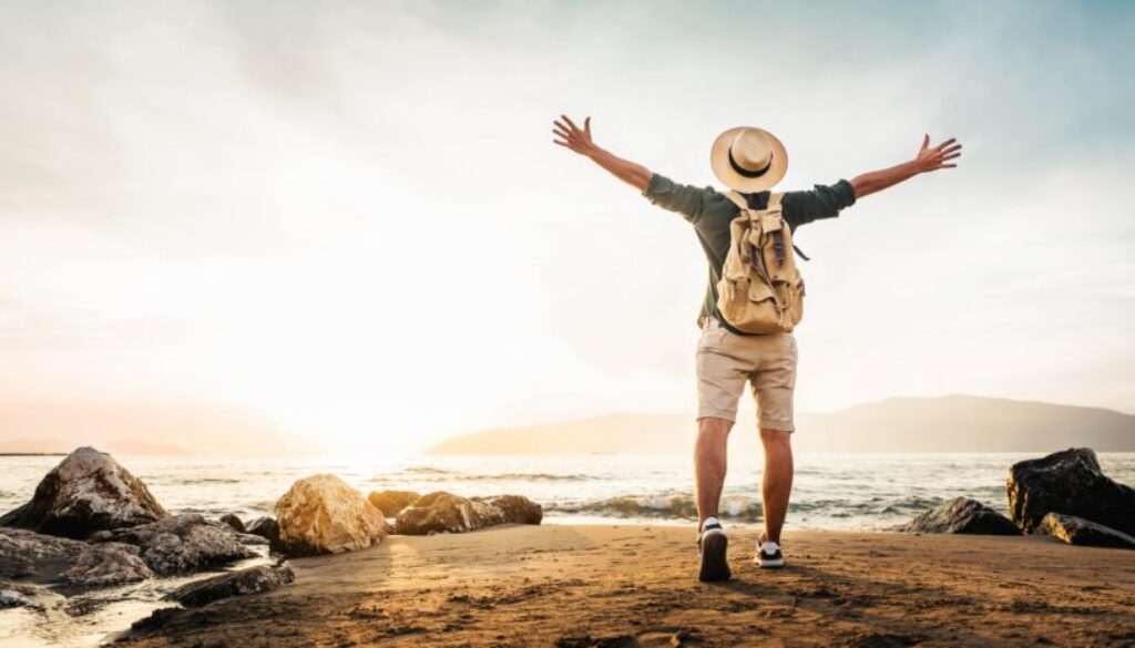 Happy man with backpack standing with arms up at the beach - Delightful tourist enjoying summer vacation by the seaside - Traveling life style and well being concept